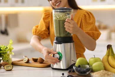 Photo of Young woman making delicious smoothie with blender at white marble table in kitchen