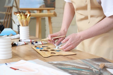 Woman with brush using palette at wooden table in drawing studio, closeup