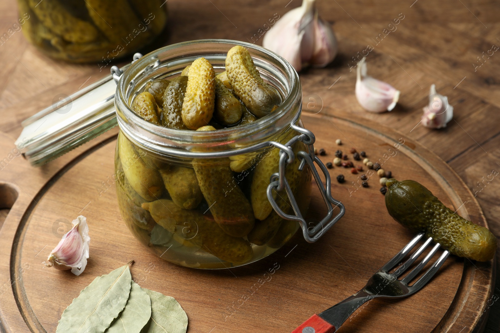 Photo of Pickled cucumbers in open jar, spices and fork on wooden table, closeup