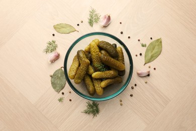 Pickled cucumbers in bowl surrounded by spices on light wooden table, flat lay