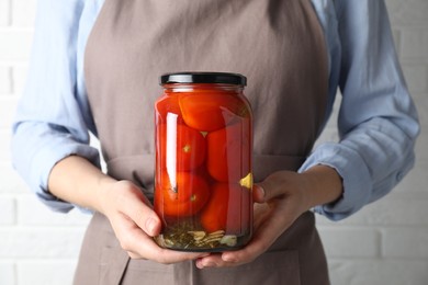 Photo of Woman holding jar with tasty pickled tomatoes against white brick wall, closeup