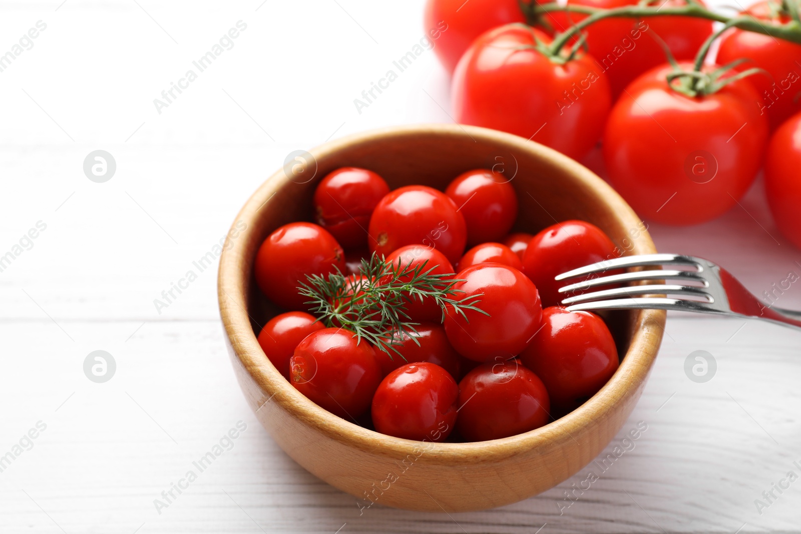 Photo of Tasty pickled tomatoes in bowl, fresh vegetables, dill and fork on white wooden table