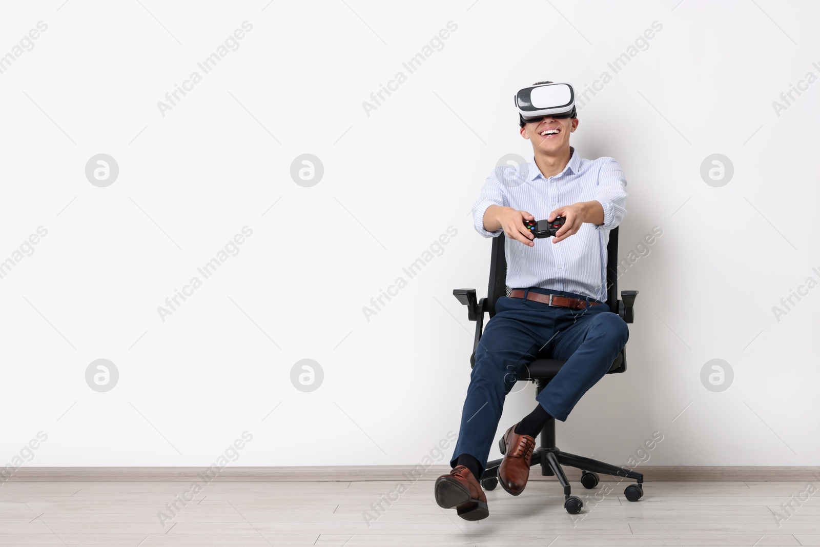 Photo of Happy young man with virtual reality headset and controller sitting on chair near white wall, space for text