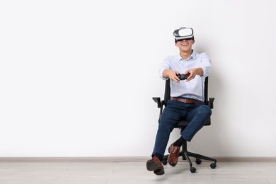 Happy young man with virtual reality headset and controller sitting on chair near white wall, space for text