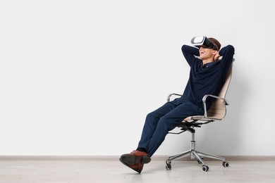 Photo of Happy young man with virtual reality headset sitting on chair near white wall, space for text