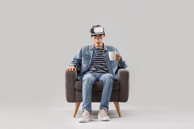 Photo of Happy young man with virtual reality headset and cup of drink sitting on armchair against light grey background