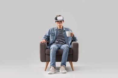 Happy young man with virtual reality headset and cup of drink sitting on armchair against light grey background