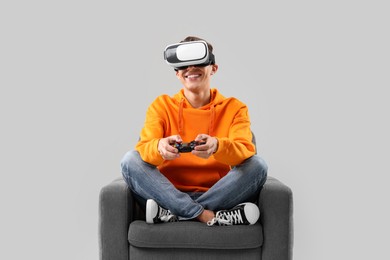 Happy young man with virtual reality headset and controller sitting on armchair against light grey background