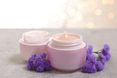 Photo of Jars of creams and ageratum flowers on gray table against light background with blurred lights, closeup