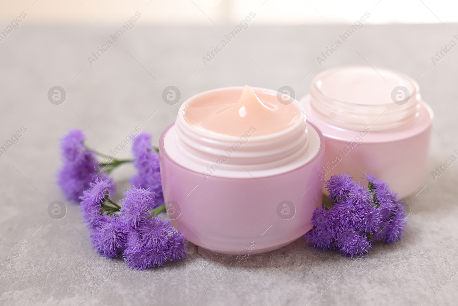 Photo of Jars of creams and ageratum flowers on gray table, closeup