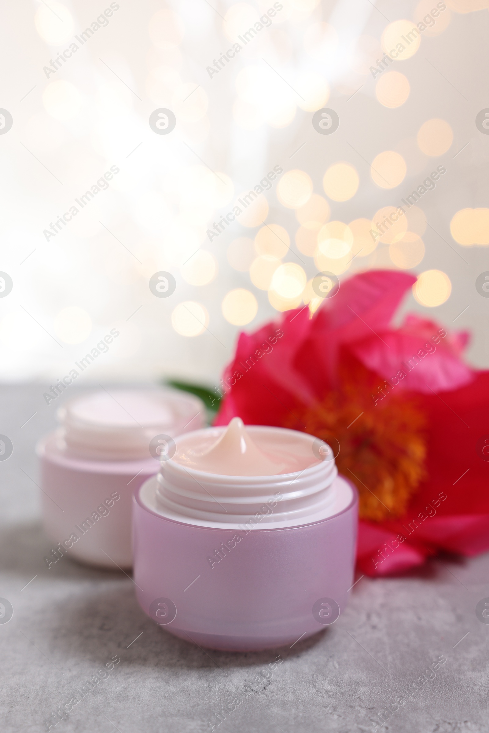 Photo of Jars of creams and peony flower on gray table against light background with blurred lights