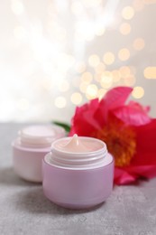 Photo of Jars of creams and peony flower on gray table against light background with blurred lights