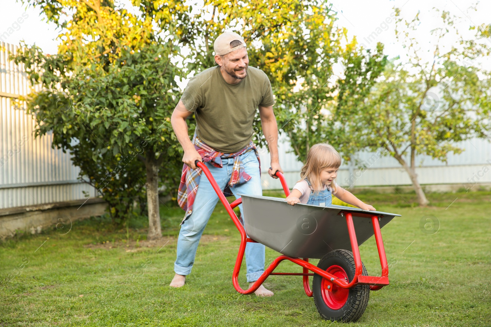 Photo of Father pushing wheelbarrow with his daughter outdoors