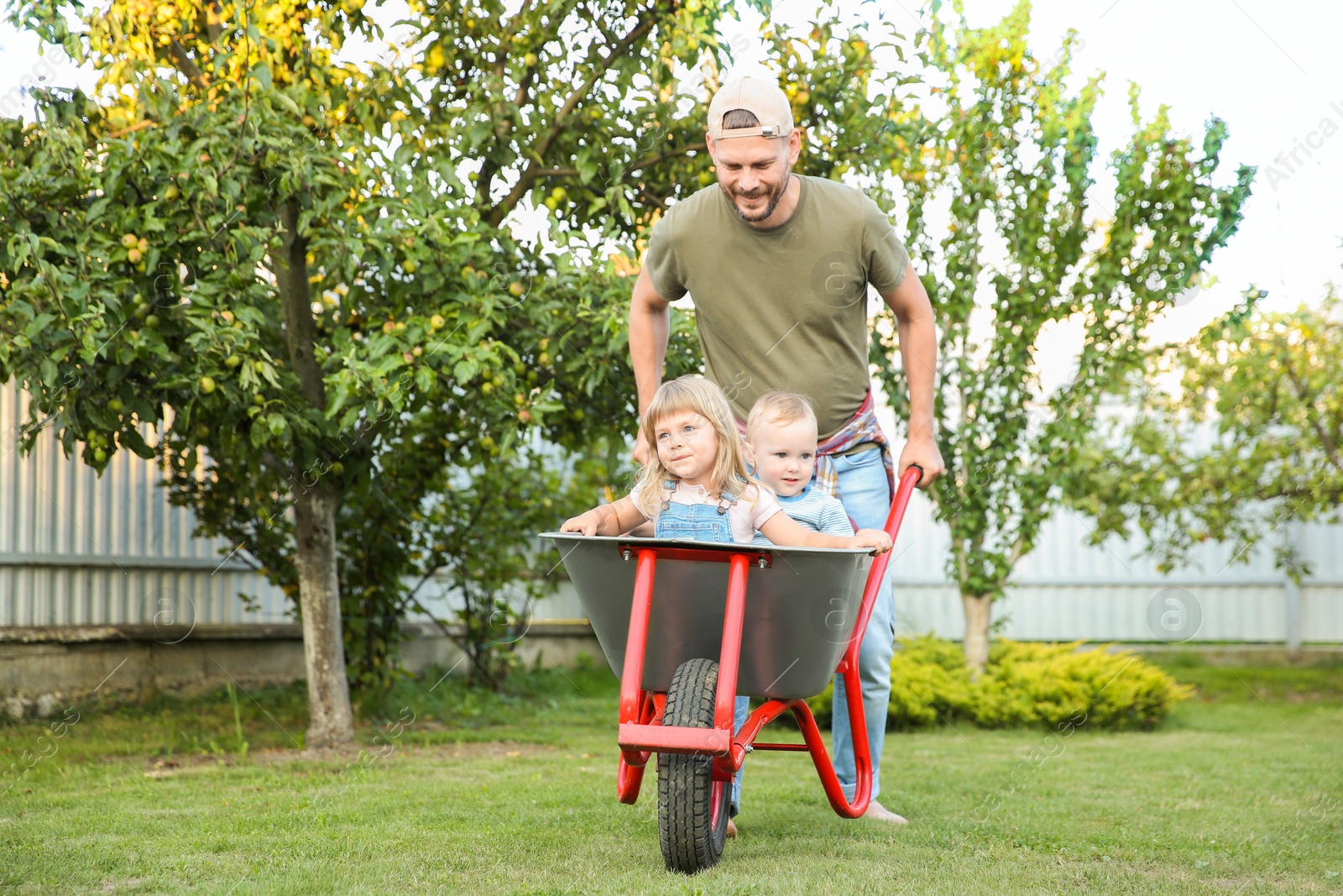 Photo of Father pushing wheelbarrow with his kids outdoors