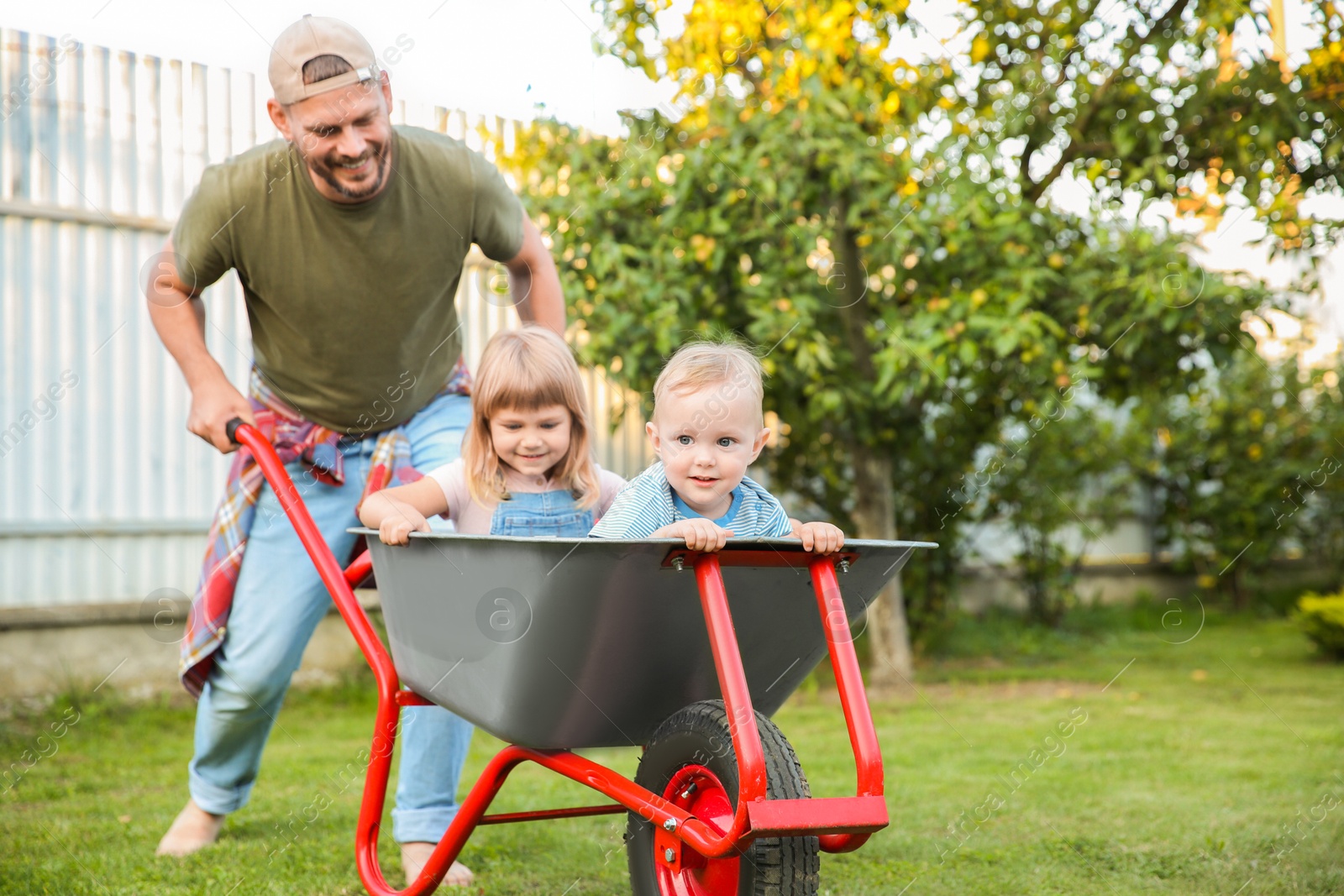 Photo of Father pushing wheelbarrow with his kids outdoors, space for text