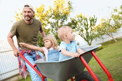 Photo of Father pushing wheelbarrow with his kids outdoors