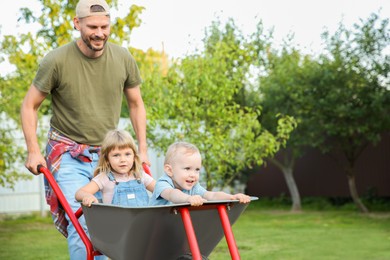 Photo of Father pushing wheelbarrow with his kids outdoors, space for text