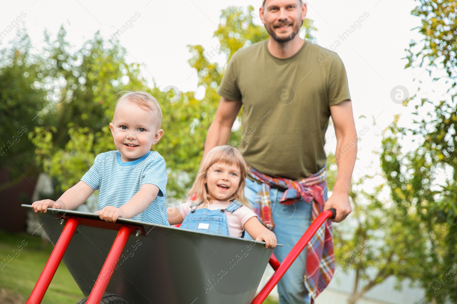 Photo of Father pushing wheelbarrow with his kids outdoors