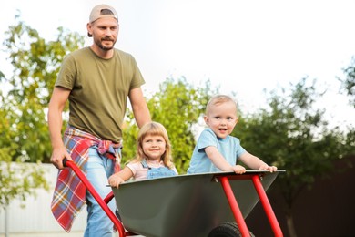 Photo of Father pushing wheelbarrow with his kids outdoors