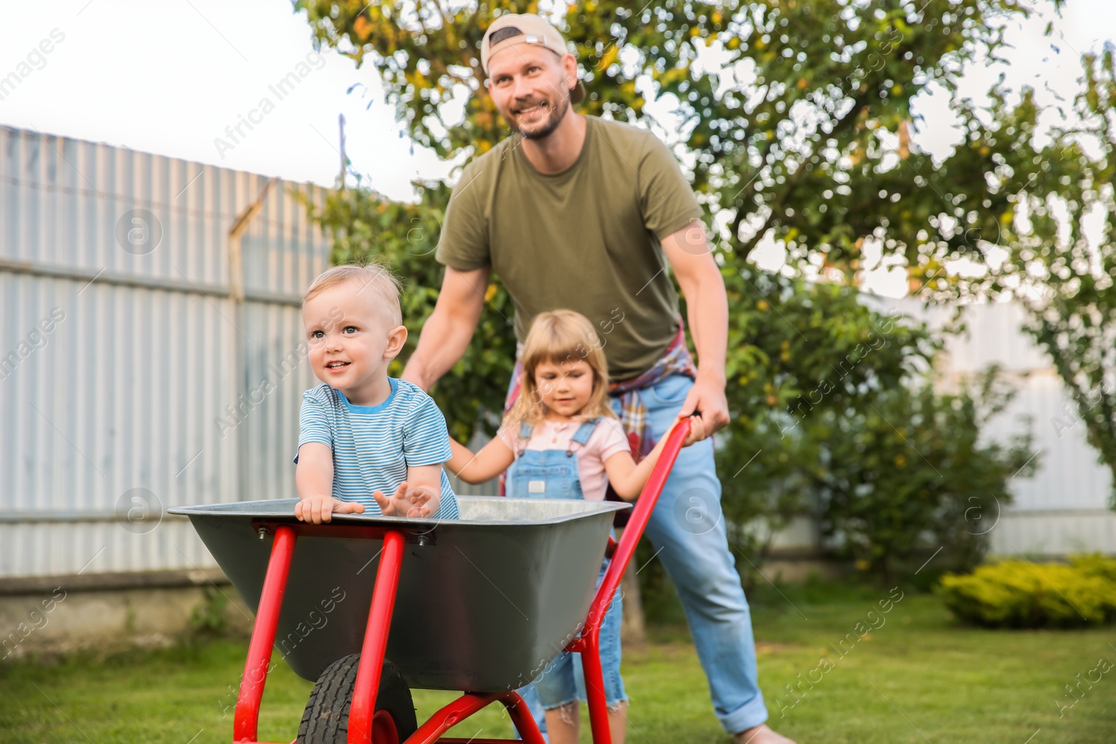 Photo of Father pushing wheelbarrow with his kids outdoors, space for text