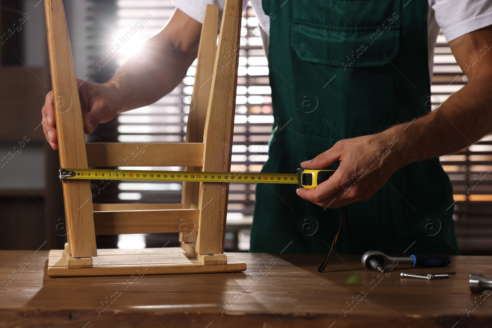 Photo of Man using tape measure while repairing wooden stool indoors, closeup