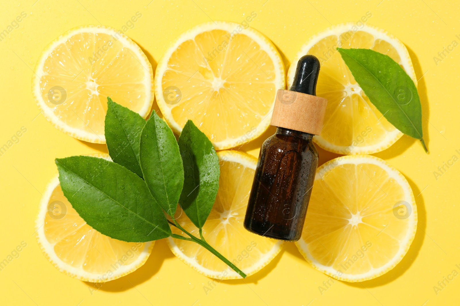 Photo of Bottle of essential oil, lemon and green leaves on yellow background, flat lay