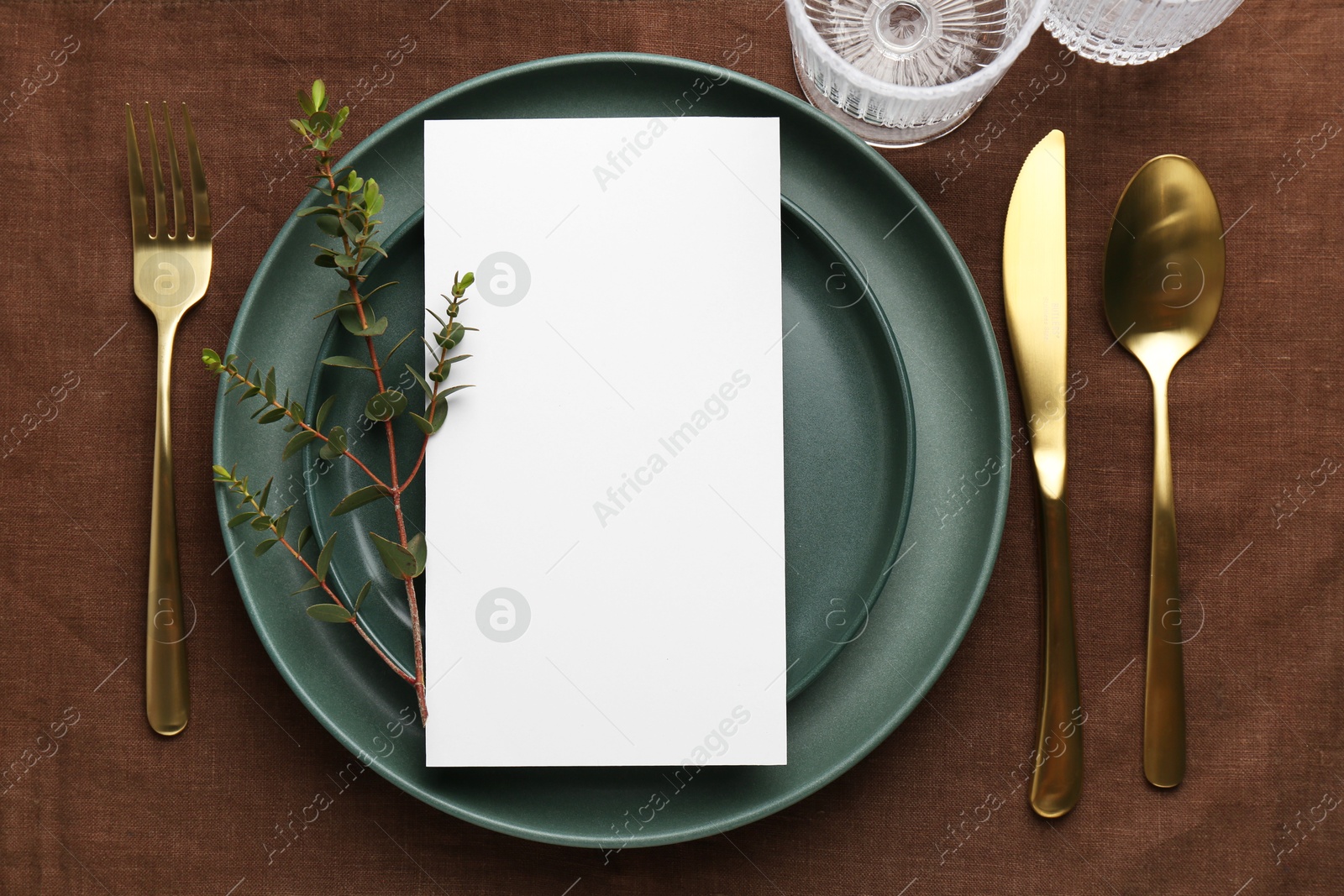 Photo of Empty menu, plates, cutlery, glass and floral decor on brown tablecloth, top view. Mockup for design