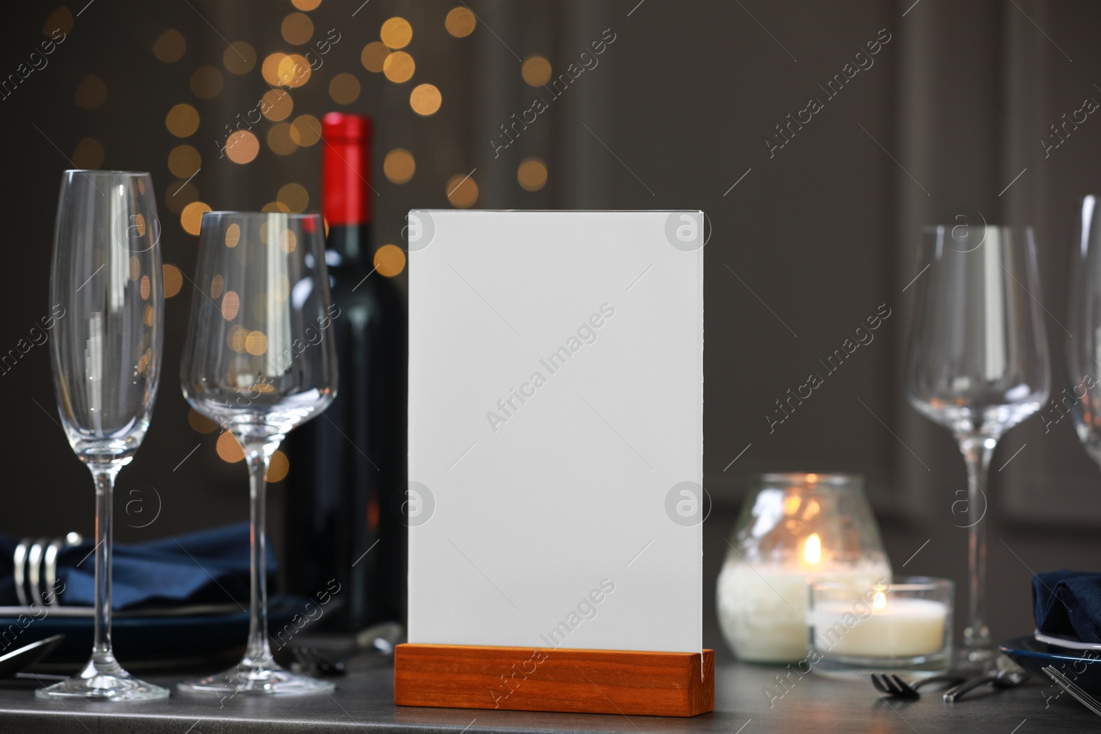 Photo of Menu holder, clean dishware and burning candles on gray table against blurred lights