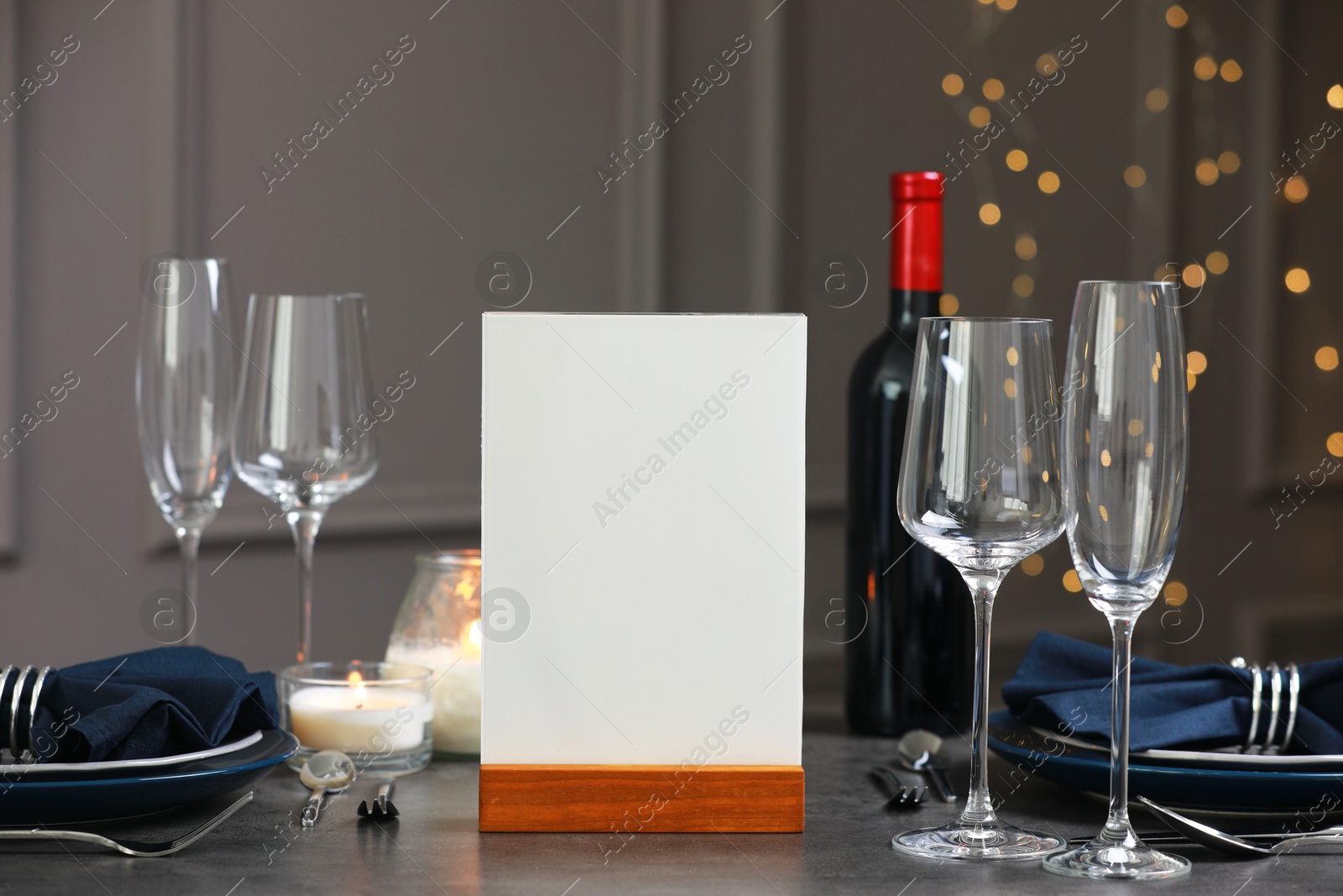 Photo of Menu holder, clean dishware and burning candles on gray table against blurred lights