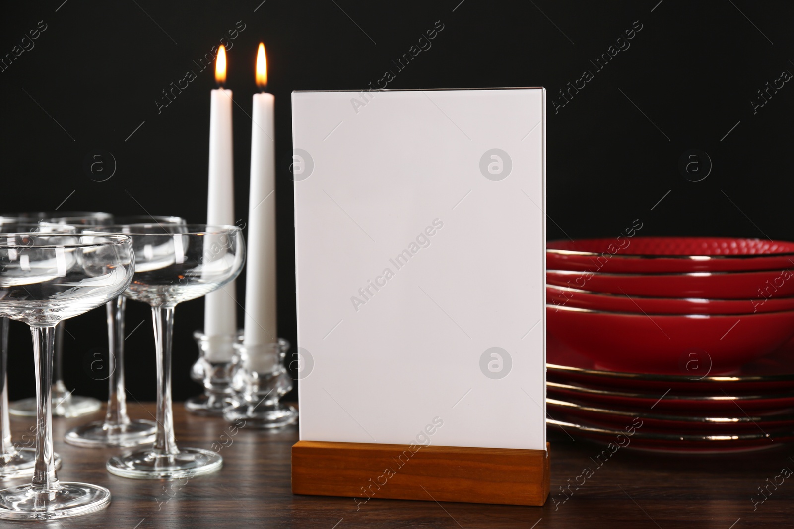 Photo of Menu holder, burning candles and clean dishware on wooden table against black background