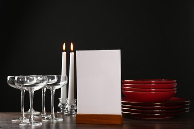 Photo of Menu holder, burning candles and clean dishware on wooden table against black background