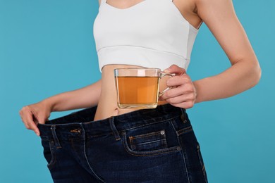 Photo of Weight loss concept. Woman with cup of diet tea and big pants on light blue background, closeup