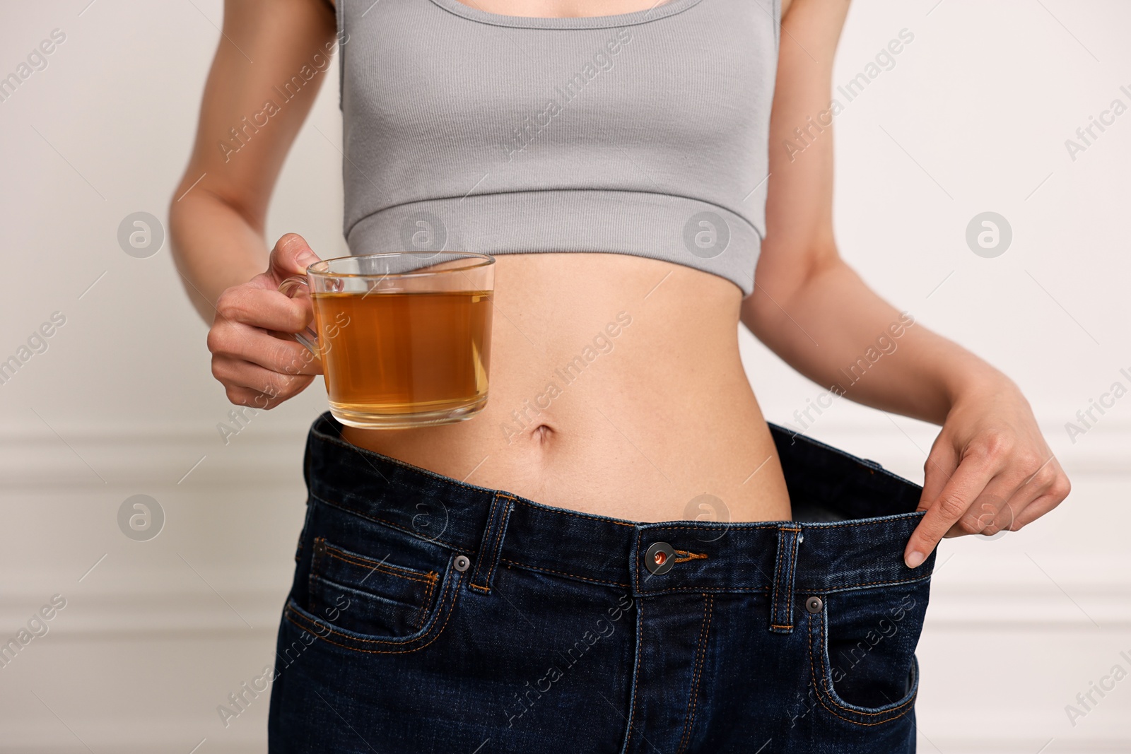 Photo of Weight loss concept. Woman with cup of diet tea and big pants near white wall, closeup