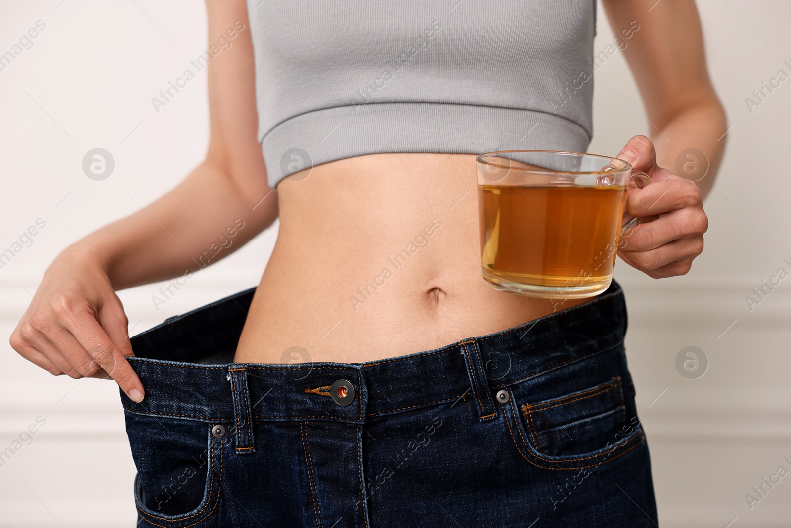 Photo of Weight loss concept. Woman with cup of diet tea and big pants near white wall, closeup