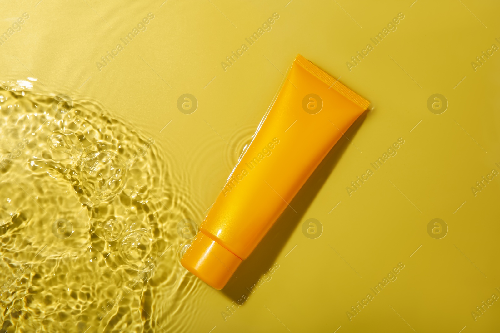 Photo of Tube with cream in water on yellow background, top view. Cosmetic product