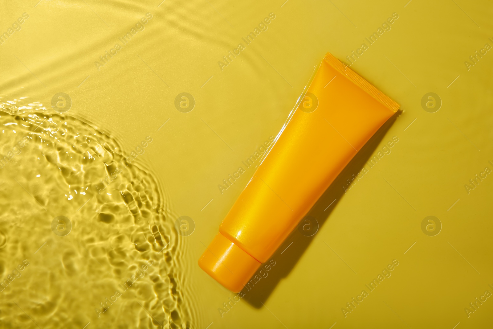 Photo of Tube with cream in water on yellow background, top view. Cosmetic product