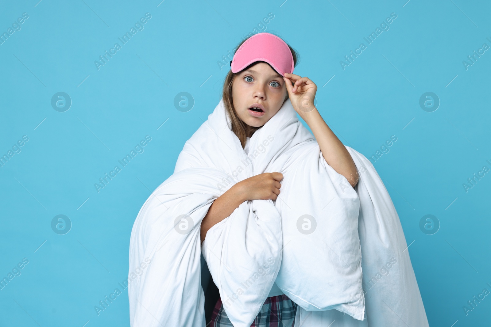 Photo of Overslept girl with sleep mask wrapped in blanket on light blue background