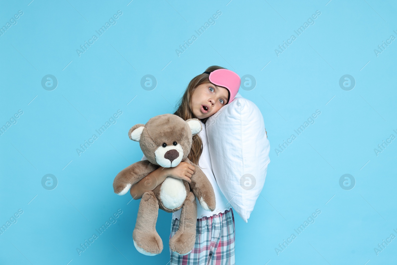 Photo of Overslept girl with sleep mask, pillow and teddy bear on light blue background