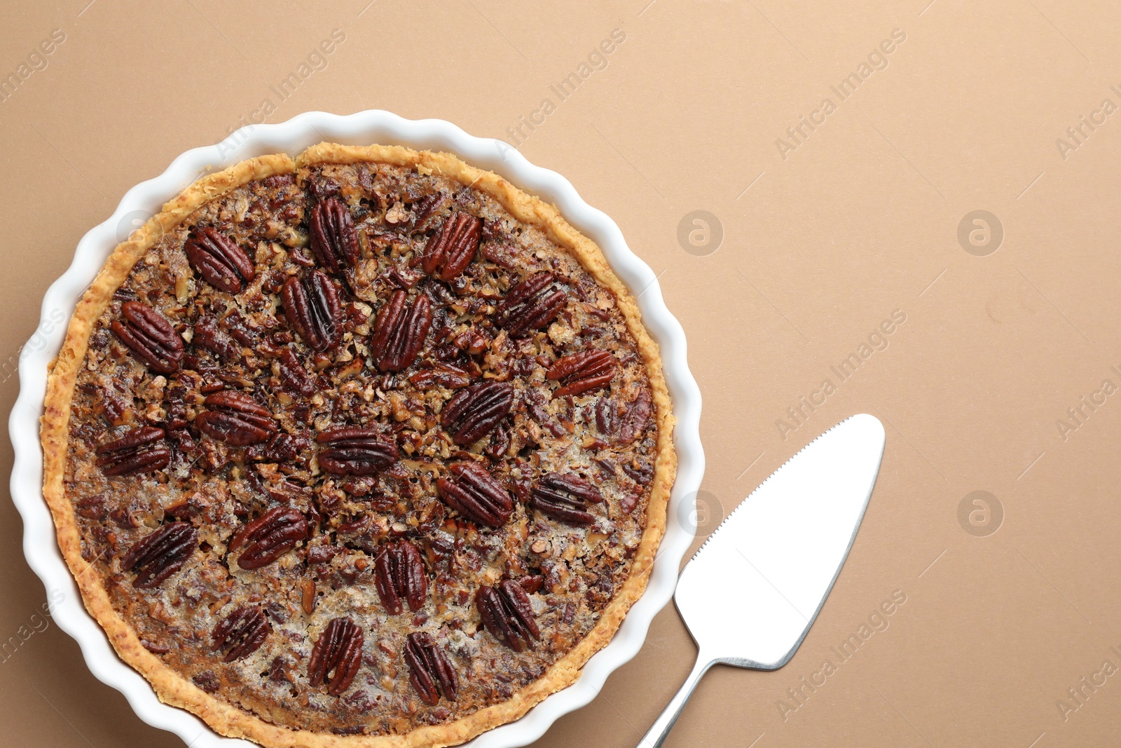 Photo of Delicious pecan pie in baking dish and cake server on light brown background, flat lay. Space for text