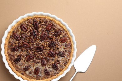 Photo of Delicious pecan pie in baking dish and cake server on light brown background, flat lay. Space for text