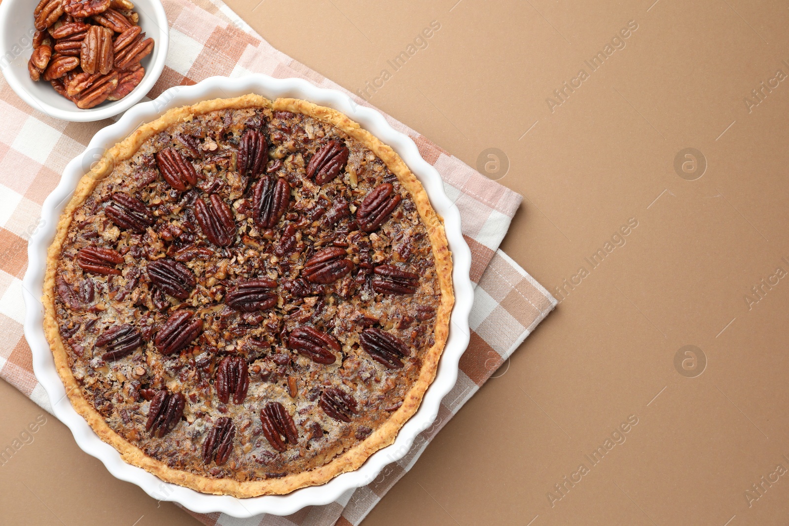 Photo of Delicious pecan pie in baking dish and fresh nuts on light brown background, flat lay. Space for text