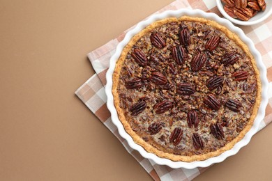 Delicious pecan pie in baking dish and fresh nuts on light brown background, flat lay. Space for text