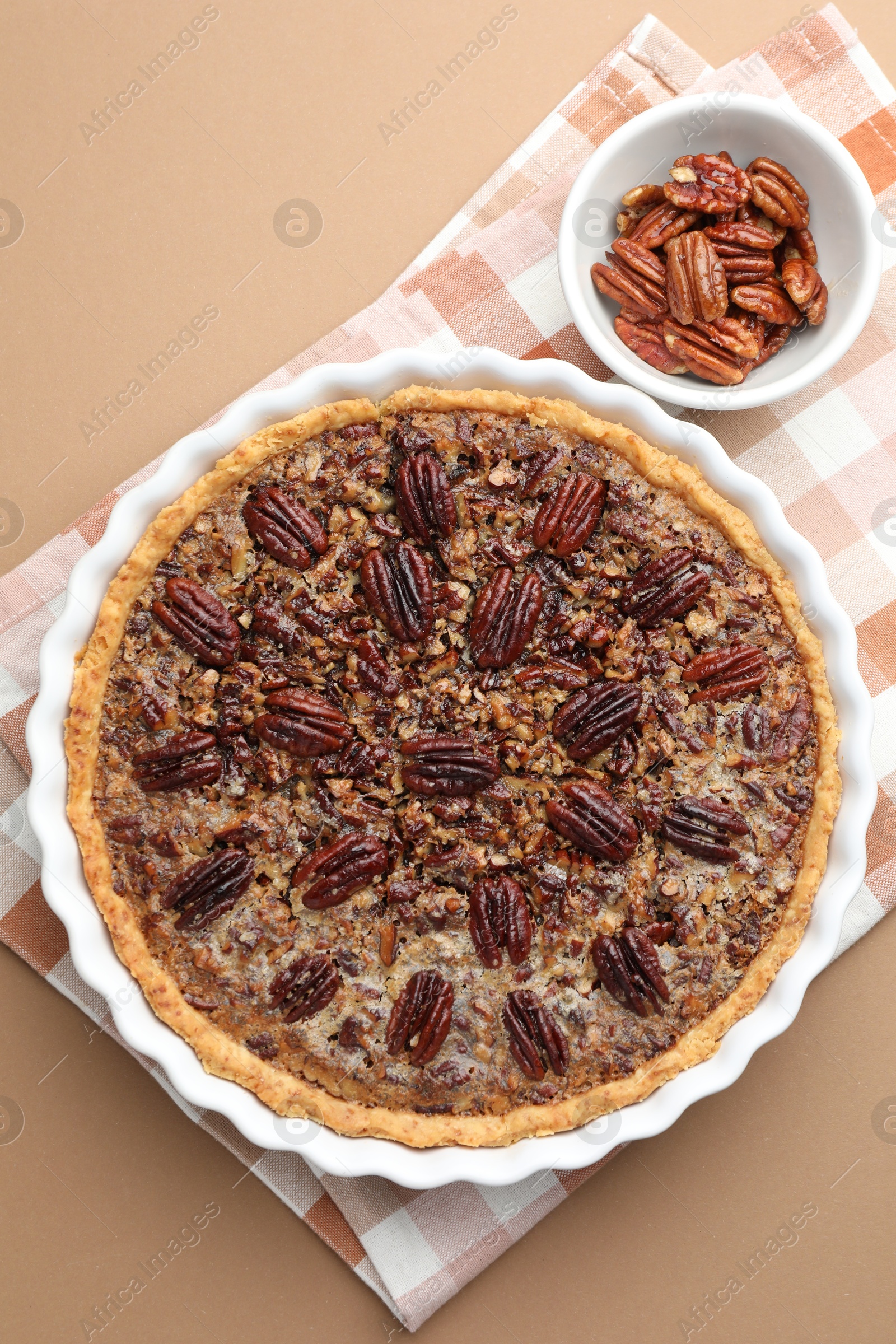 Photo of Delicious pecan pie in baking dish and bowl with fresh nuts on light brown background, flat lay