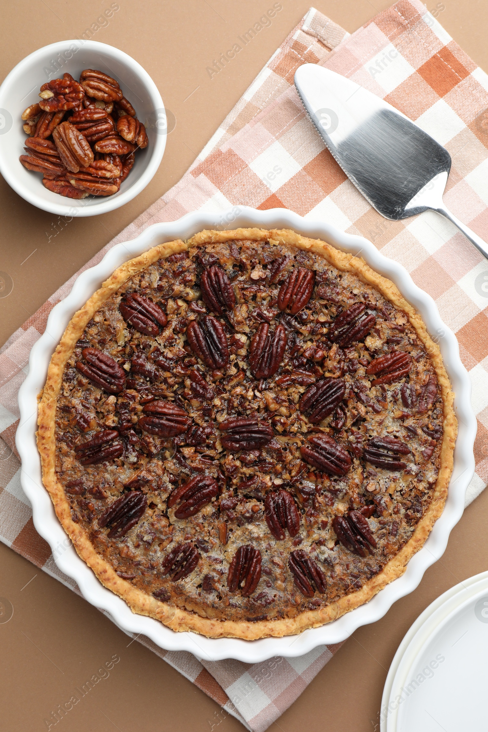 Photo of Delicious pecan pie in baking dish, bowl with fresh nuts and cake server on light brown background, flat lay