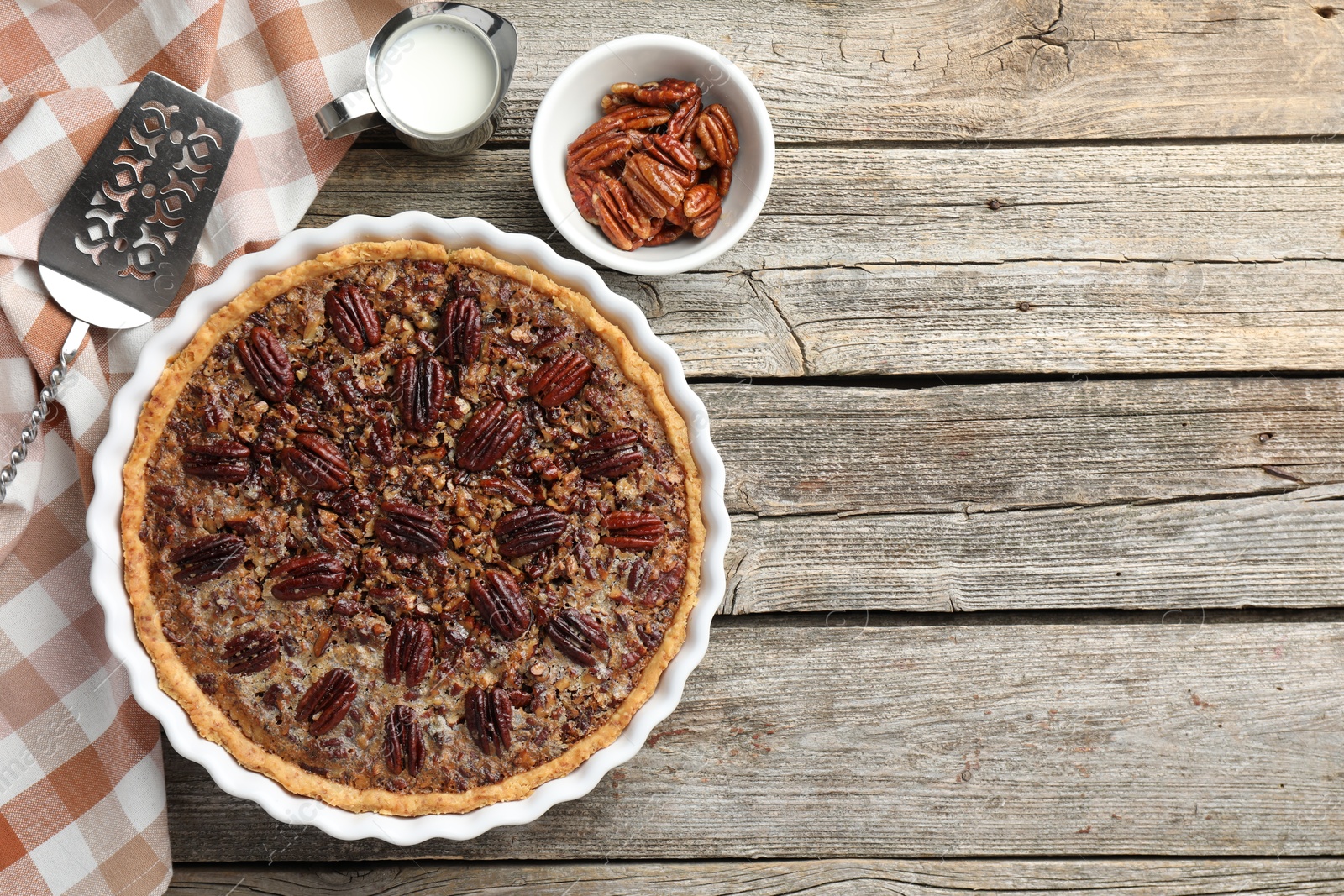 Photo of Delicious pecan pie in baking dish, fresh nuts, milk and cake server on wooden table, flat lay. Space for text