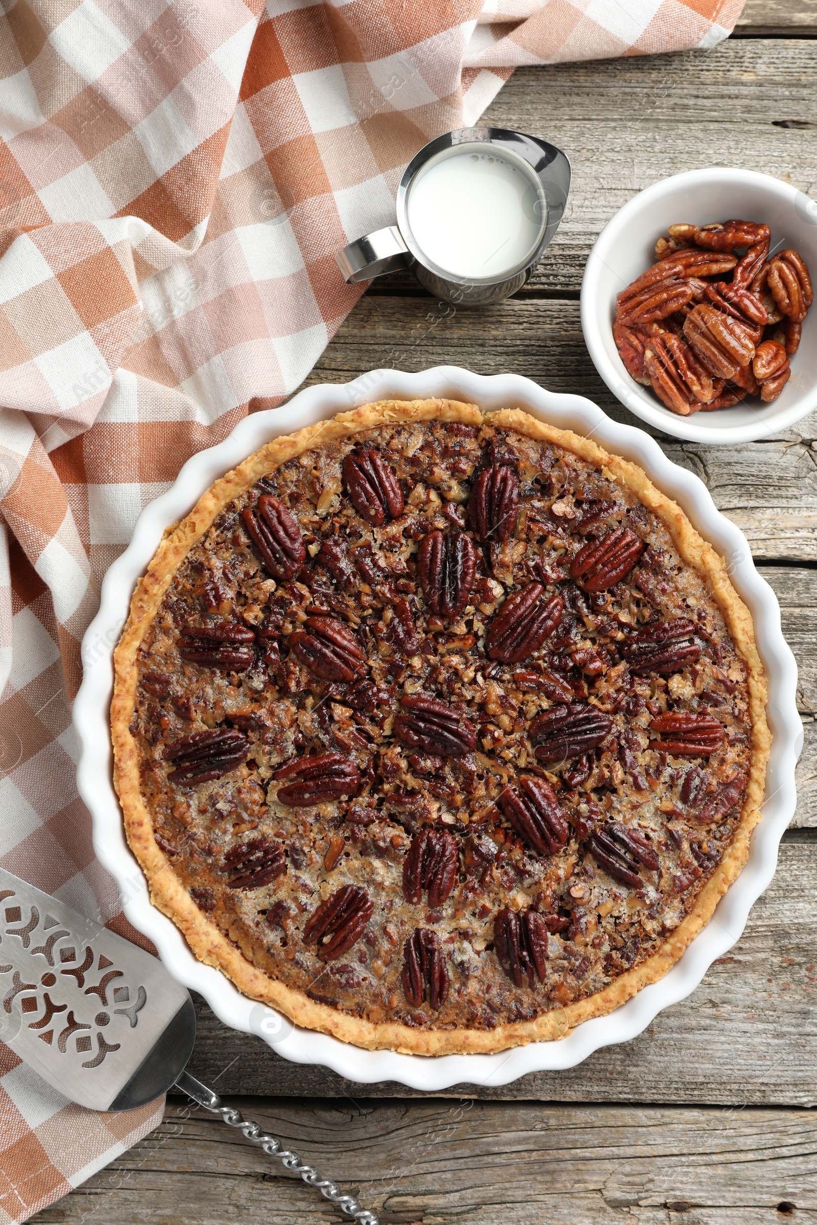 Photo of Delicious pecan pie in baking dish, fresh nuts, milk and cake server on wooden table, flat lay