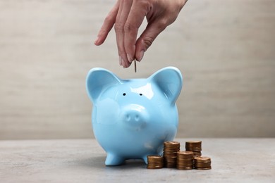 Woman putting coin into light blue piggy bank at grey table, closeup