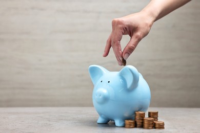 Photo of Woman putting coin into light blue piggy bank at grey table, closeup. Space for text