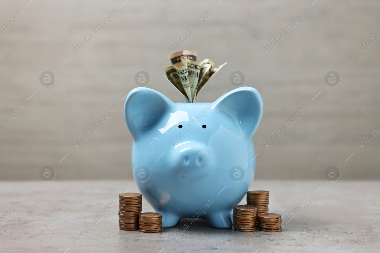 Photo of Light blue piggy bank with dollar banknotes and stacks of coins on grey table