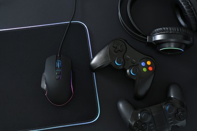 Photo of Computer mouse, mousepad, headset and game controllers on black background, flat lay